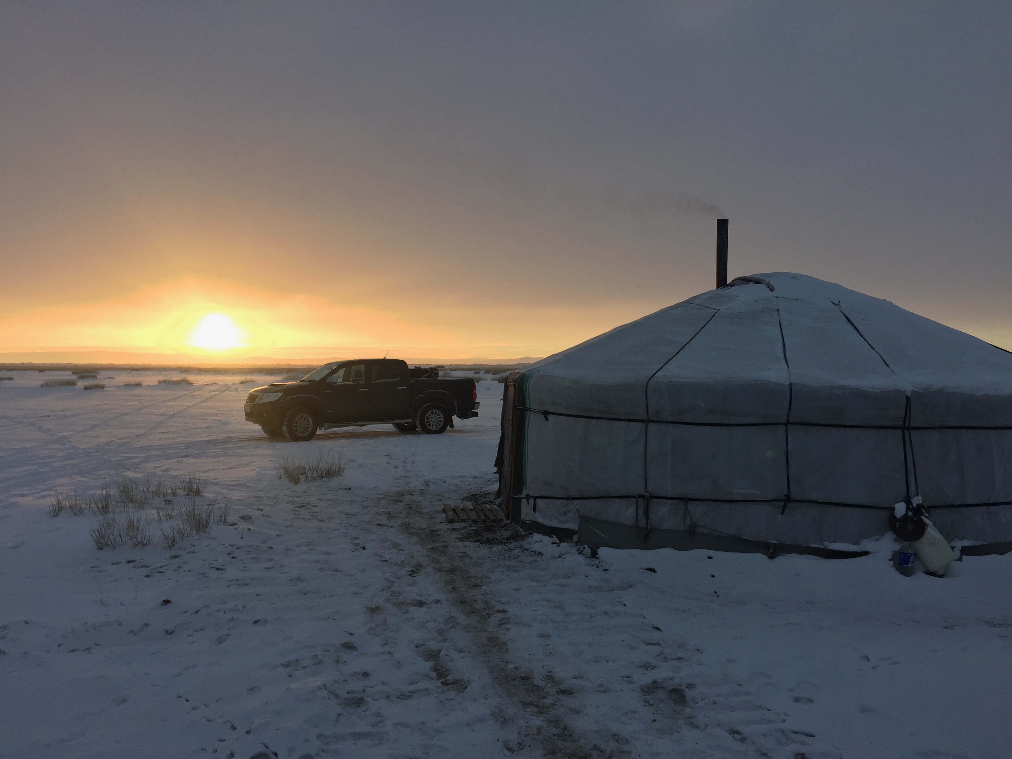 Pickup truck parked beside a yurt at sunrise on the Mongolian steppe, fresh snow on the ground