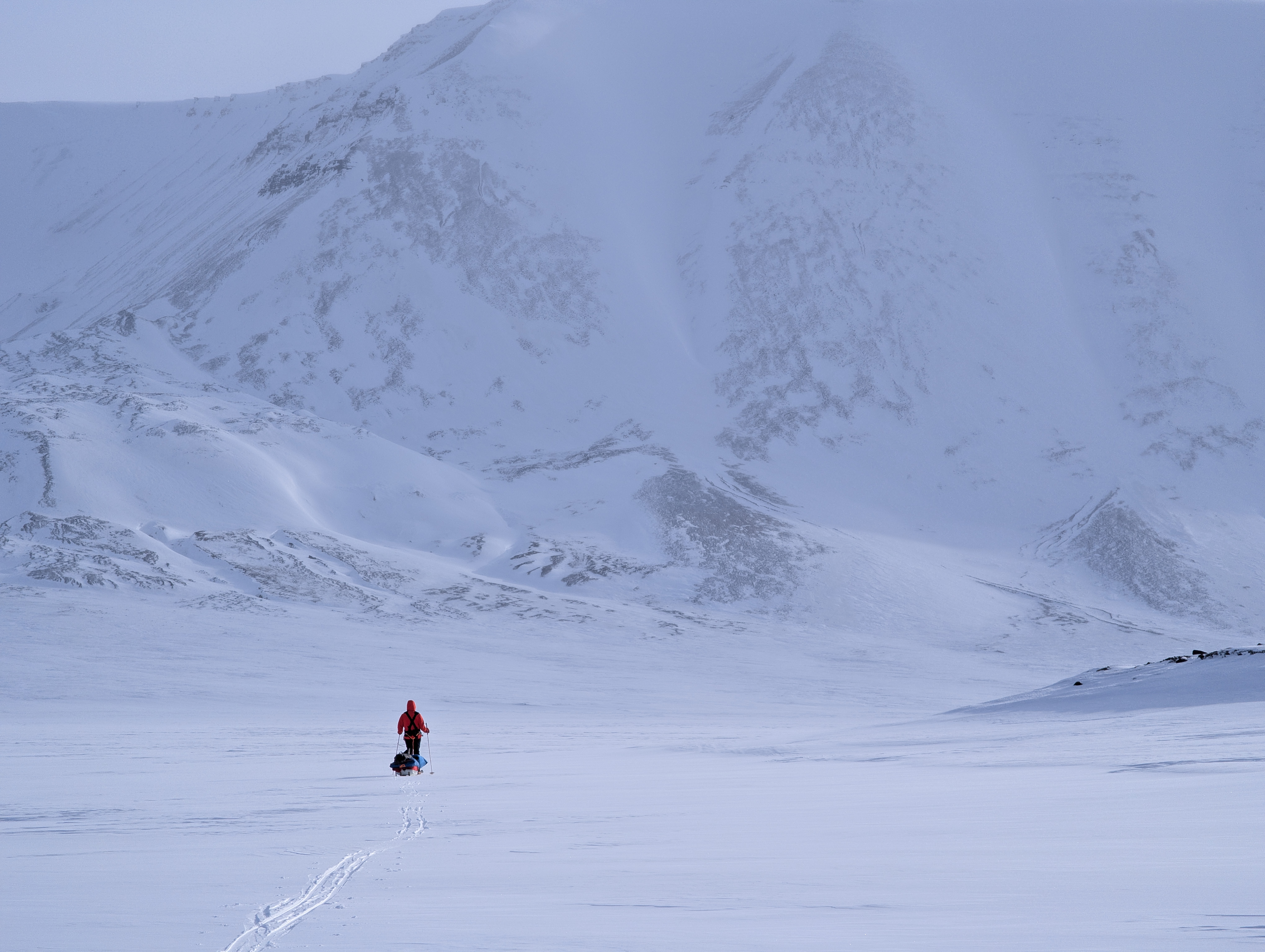 Lone figure in a red parka hauling a pulk sled toward a vast mountain ridge — Svalbard 2026